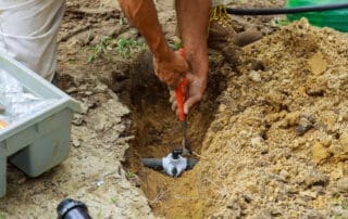 A worker kneels beside a dug-out trench and repairs an underground pipe joint, with loose soil and tools nearby.